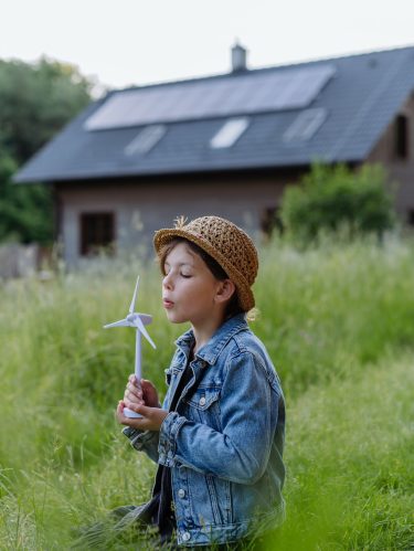Little girl with model of wind turbine, standing in front of their house with solar panels, concept